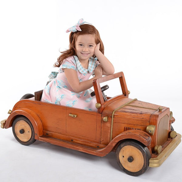 Little girl wearing Lil' Lou Lou Melly Dress in pink Classic Truck print sitting in a vintage wooden toy car