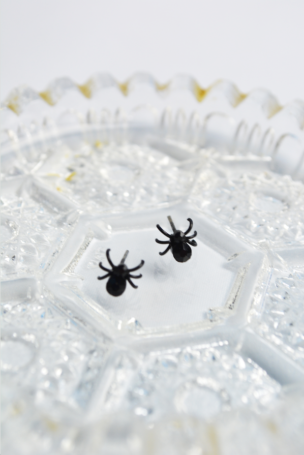 Black spider stud earrings with faceted crystal accents displayed on a vintage glass dish.