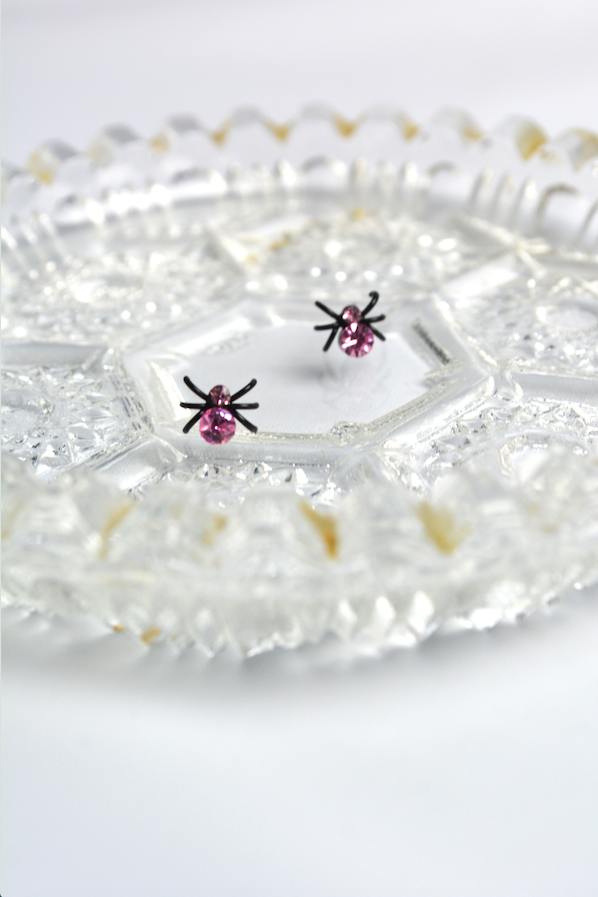 Miniature black spider stud earrings with pink faceted crystal accents displayed on a decorative vintage glass tray.
