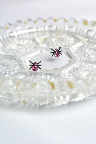 Miniature black spider stud earrings with pink faceted crystal accents displayed on a decorative vintage glass tray.