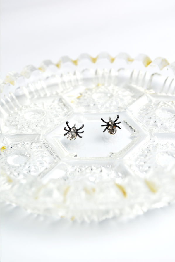 Miniature black spider stud earrings with clear faceted crystal accents displayed on a decorative vintage glass tray.
