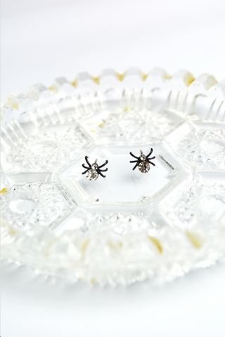 Miniature black spider stud earrings with clear faceted crystal accents displayed on a decorative vintage glass tray.
