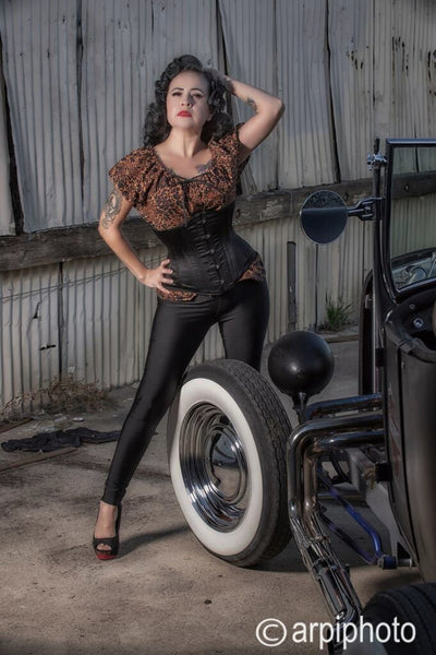 Model wearing Lil' Lou Lou's Leopard Mary Blouse styled with a waist cincher and black cigarette pants beside a vintage hot rod.