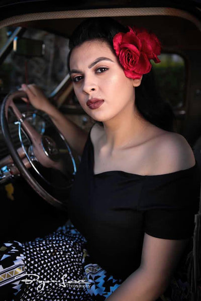 Model wearing the Rockin' Cori black off-the-shoulder top styled with a red hair flower and boogie woogie skirt while seated in a vintage car.