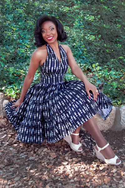 Model seated outdoors wearing the Wanda Dress in black arrow print featuring a halter neckline and full gathered swing skirt.