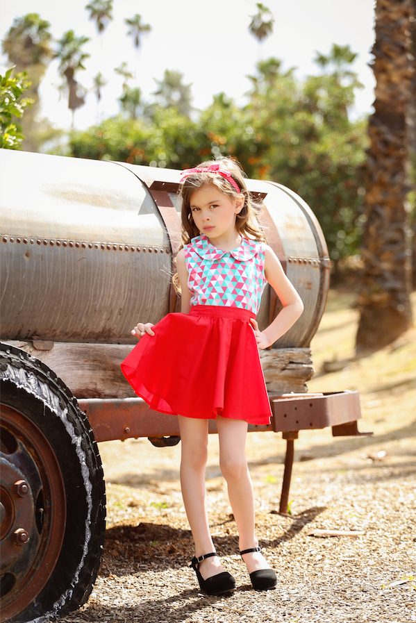 Girl wearing the Triangle Aqua Wendy Top with geometric print and Peter Pan collar paired with a red Kids Jivin' Skirt beside a vintage trailer.
